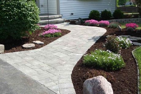 A brick walkway leading to a house surrounded by flowers and rocks.