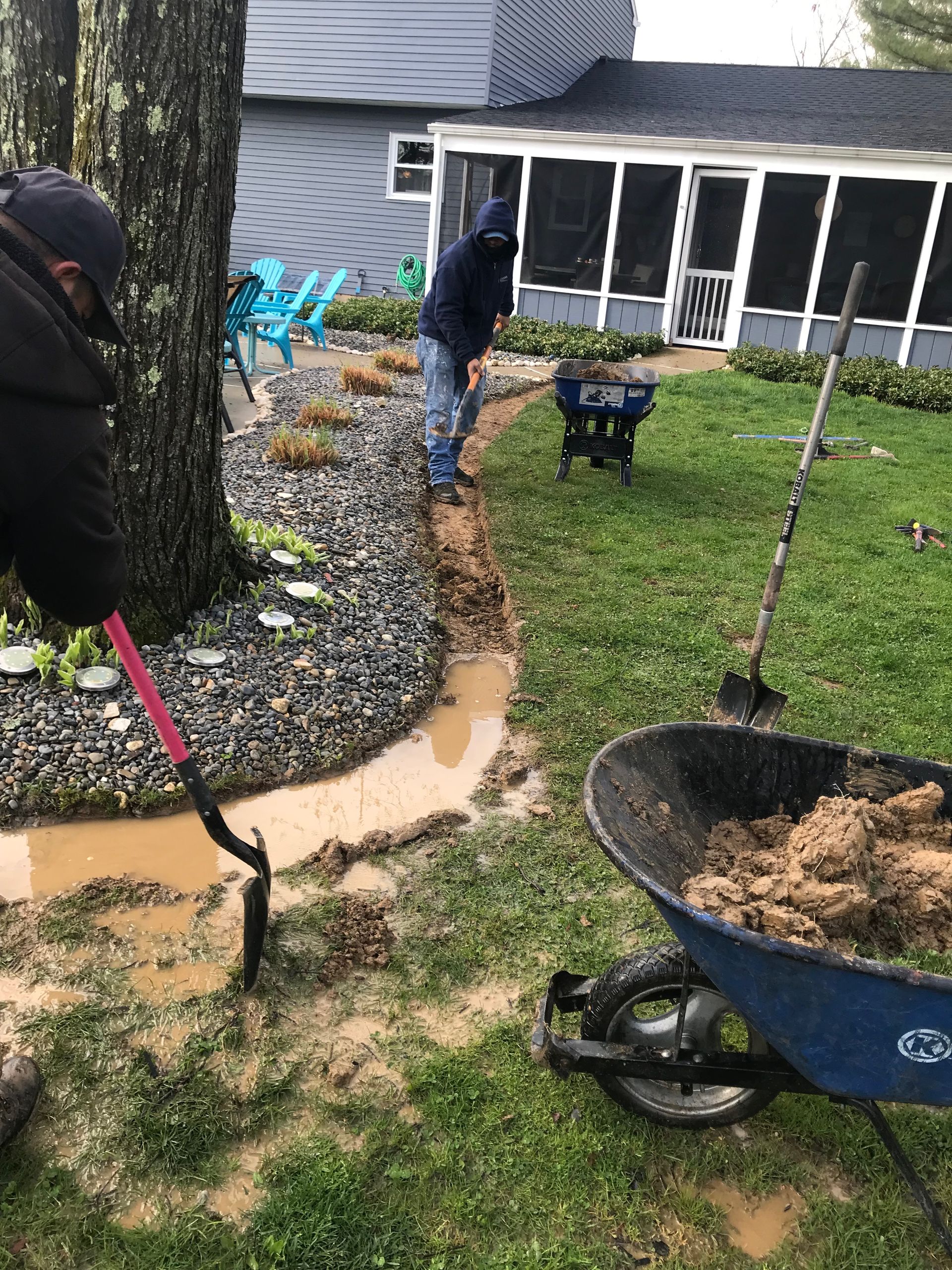 A man is digging in a muddy yard next to a wheelbarrow.