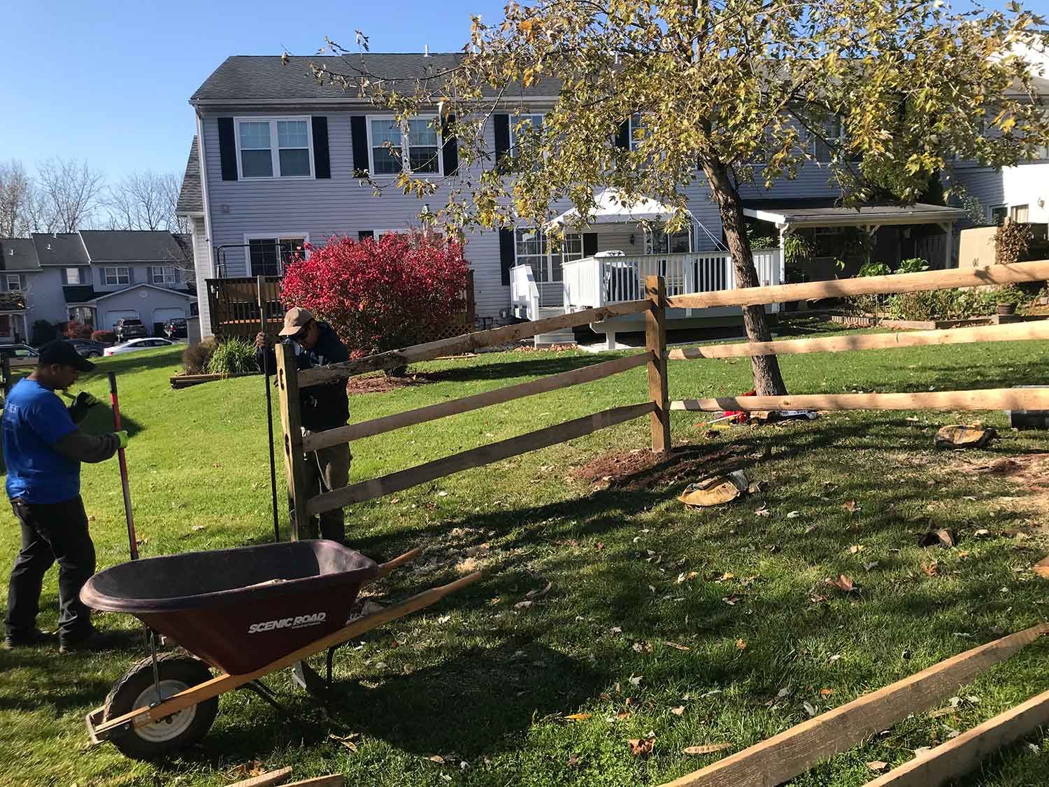 A man is pushing a wheelbarrow in front of a wooden fence.