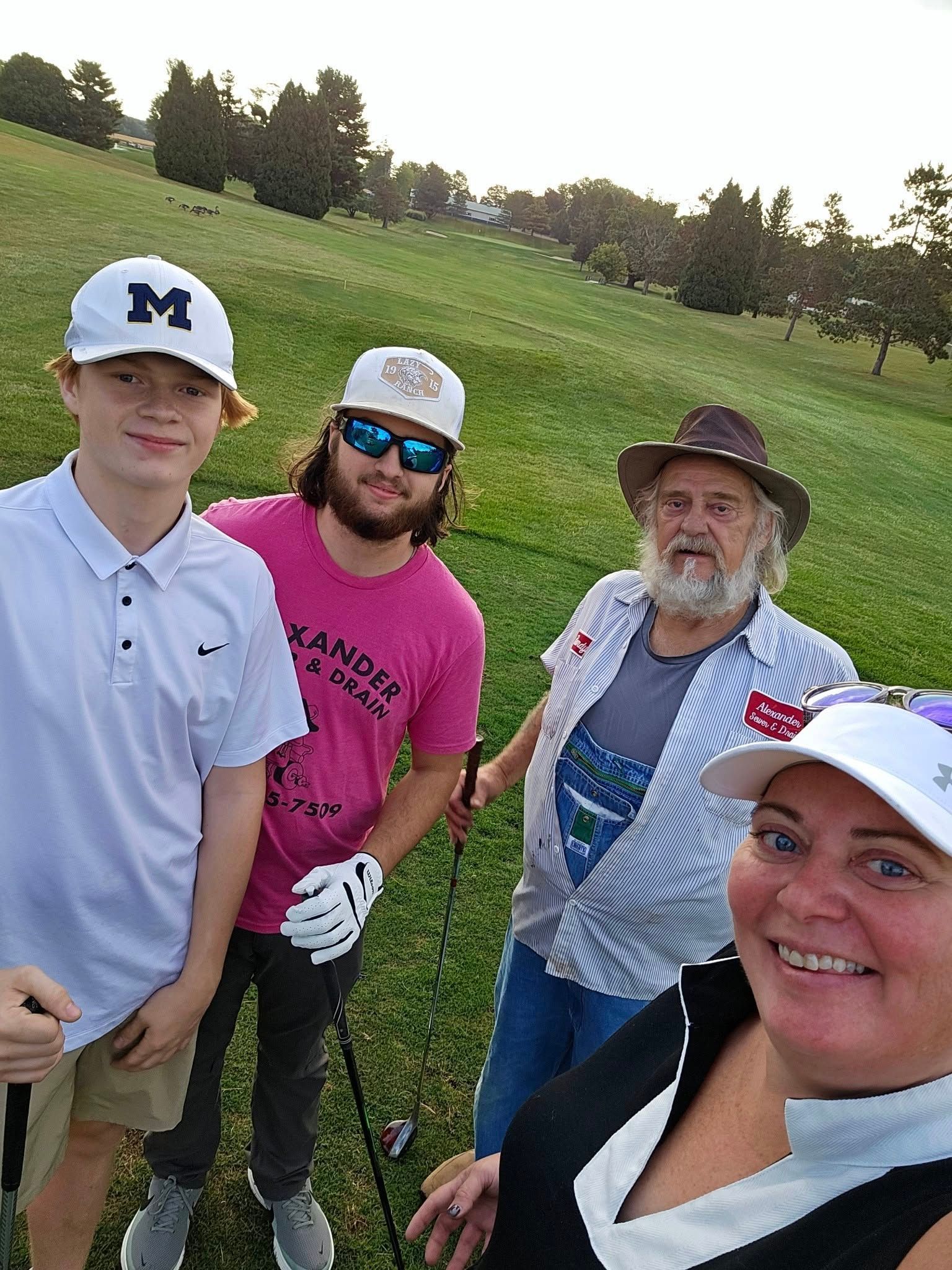 Four people posing on a golf course. Three men and a woman smile for a selfie. Green grass, blue sky.