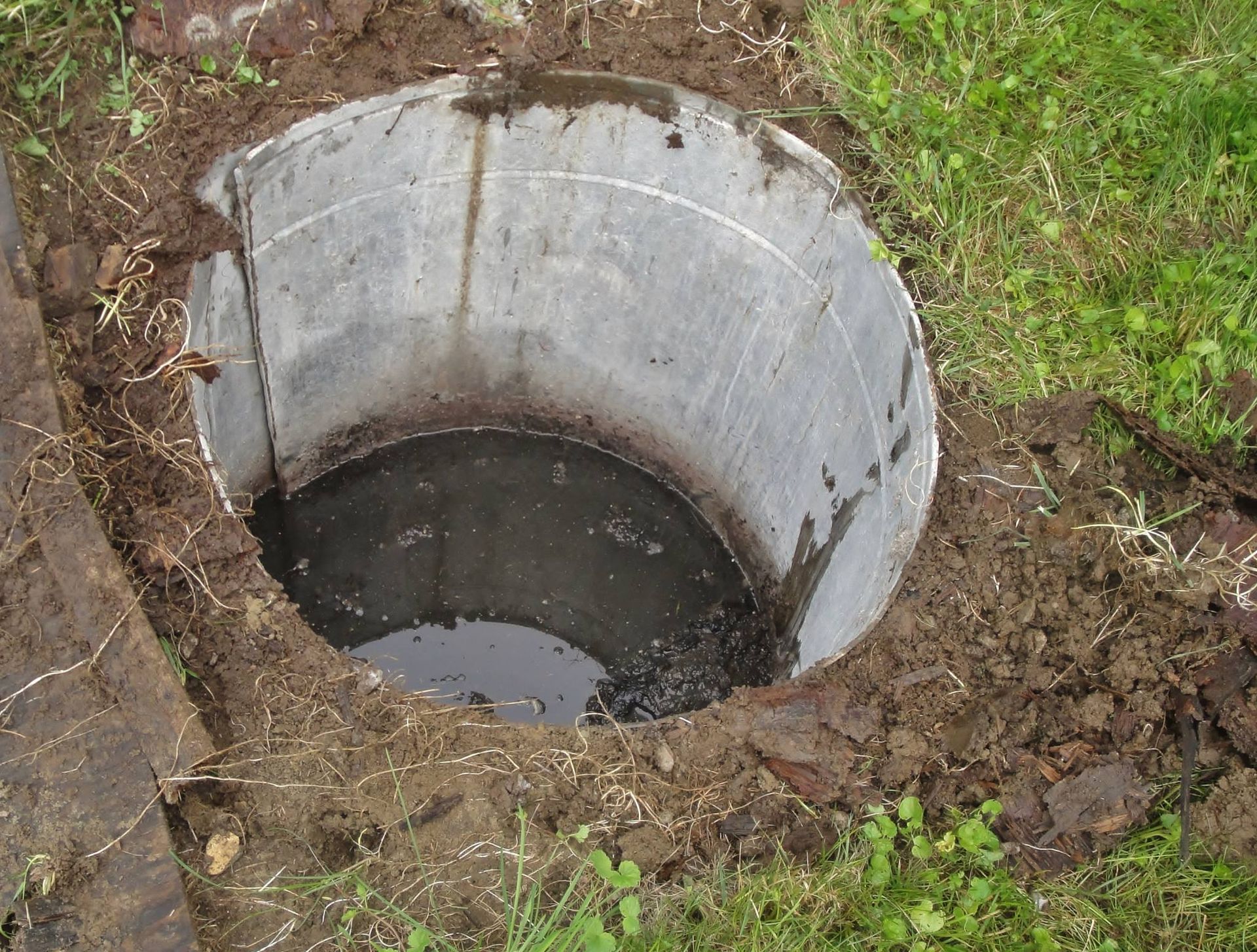 Open concrete well in dirt with water at the bottom, surrounded by grass.