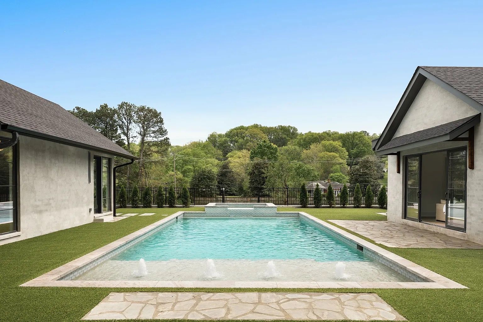 A rectangular pool between two houses with green grass and a view of trees under a blue sky.