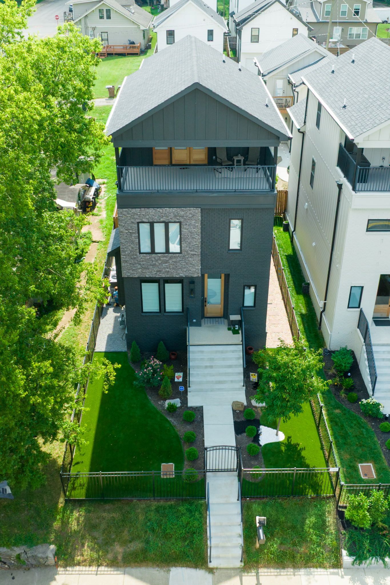 Black modern three-story house with a long walkway, flanked by trees and grass, on a sunny day.