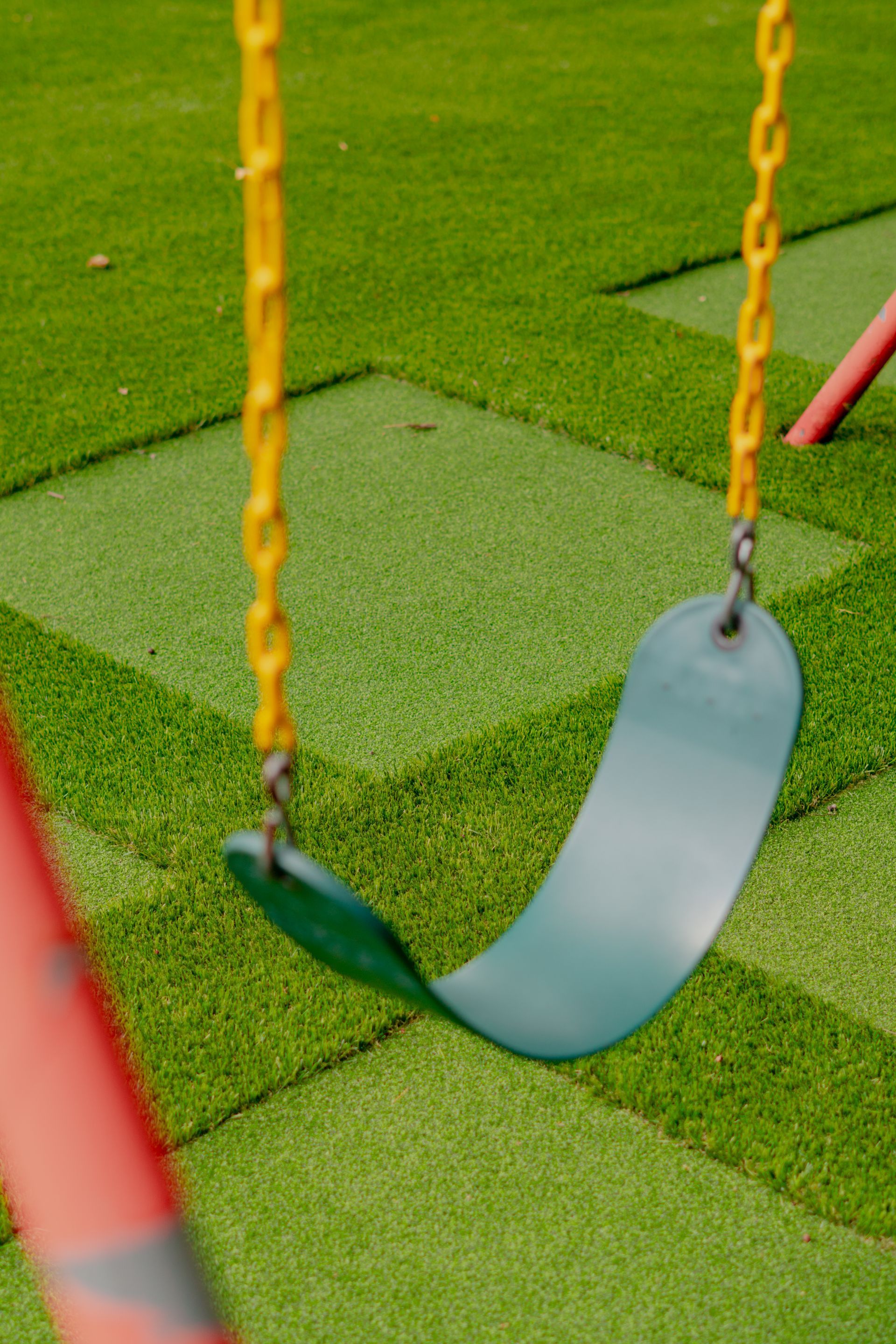 Green swing with yellow chains on a green playground surface, red support structure in the foreground.