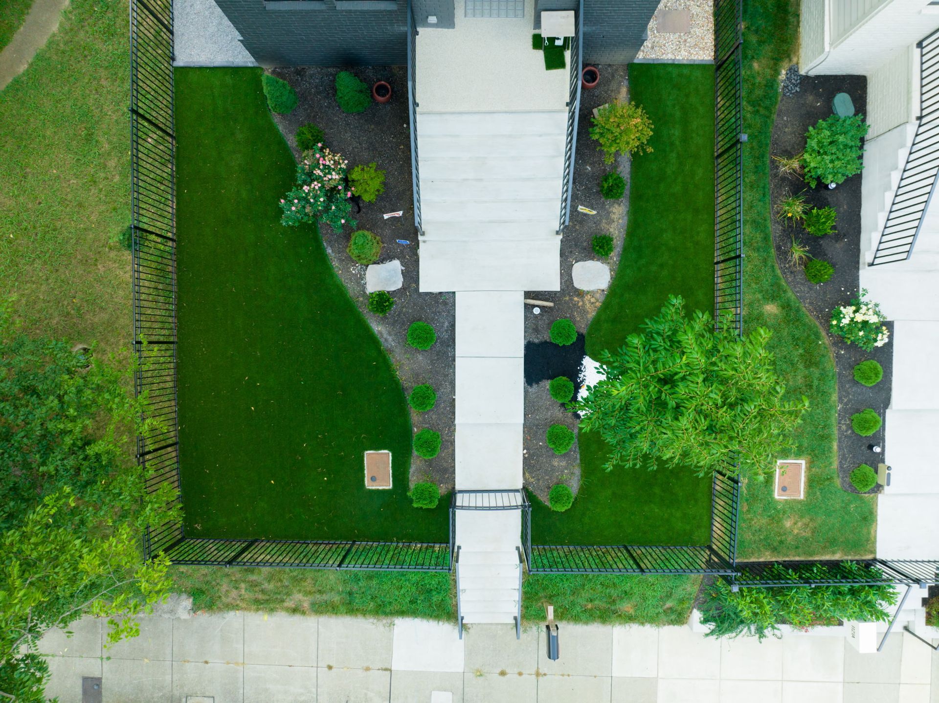 Overhead view of a house with a symmetrical yard. Lush green lawn, black fence, and gray concrete pathway.