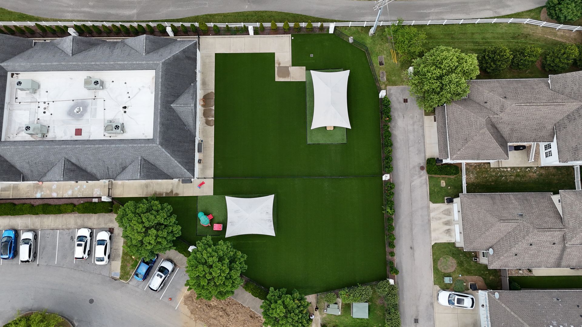 Overhead view of a green lawn area with white tents, between buildings and a parking lot.