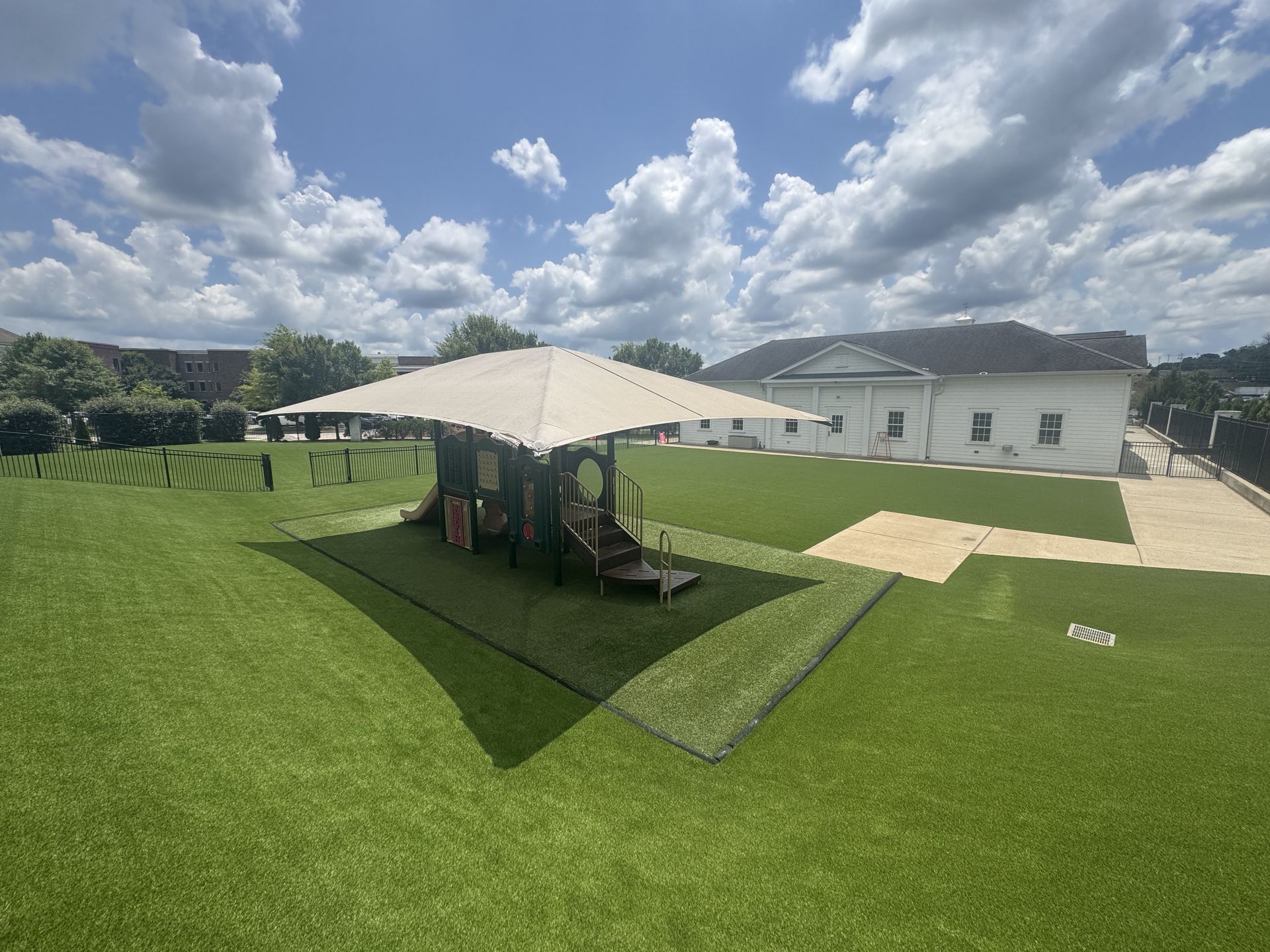 Playground under a canopy on a grassy lawn, with a white building in the background under a blue sky.