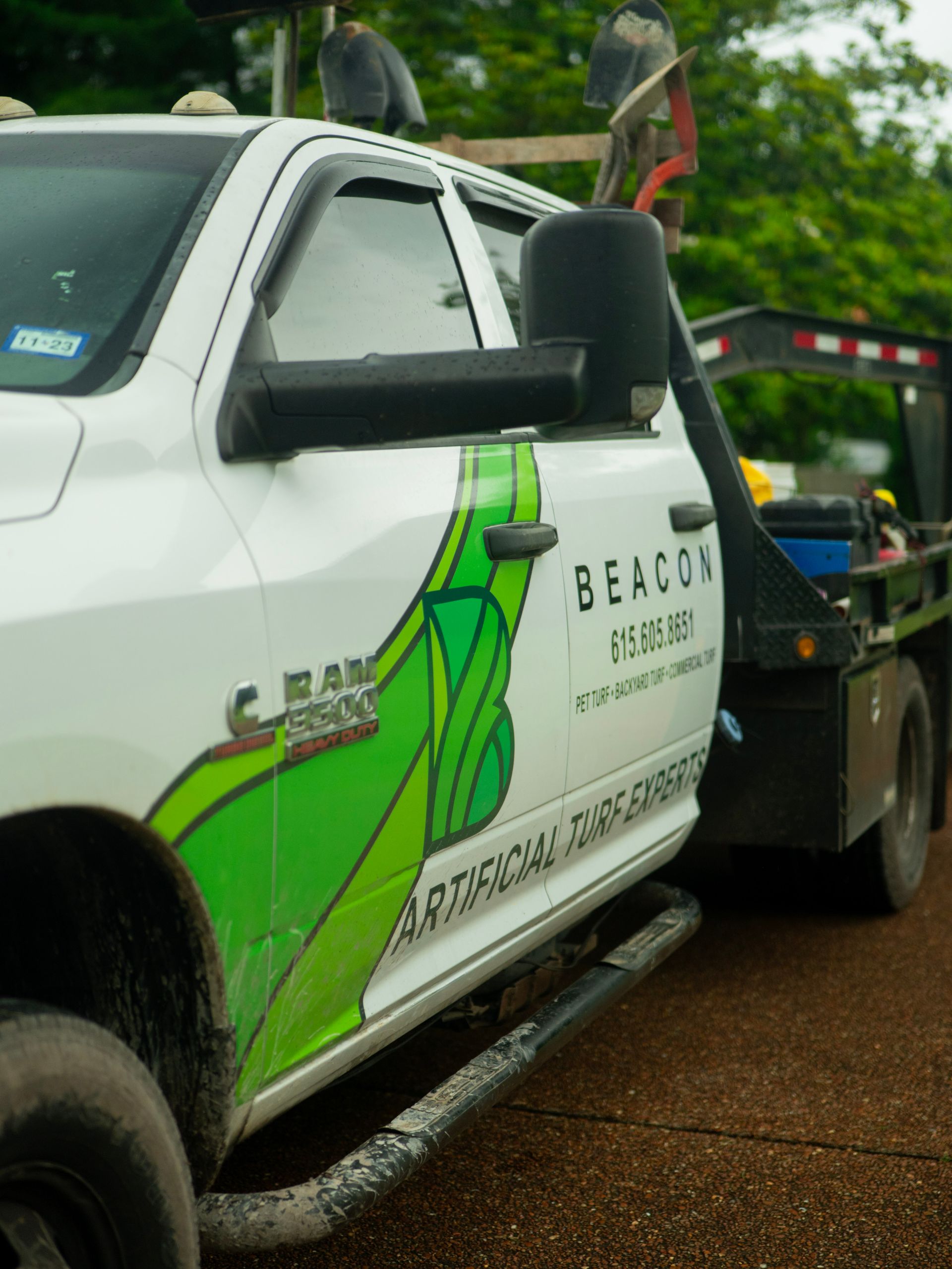 White truck with green logo for Beacon Artificial Turf Solutions, towing a flatbed trailer.