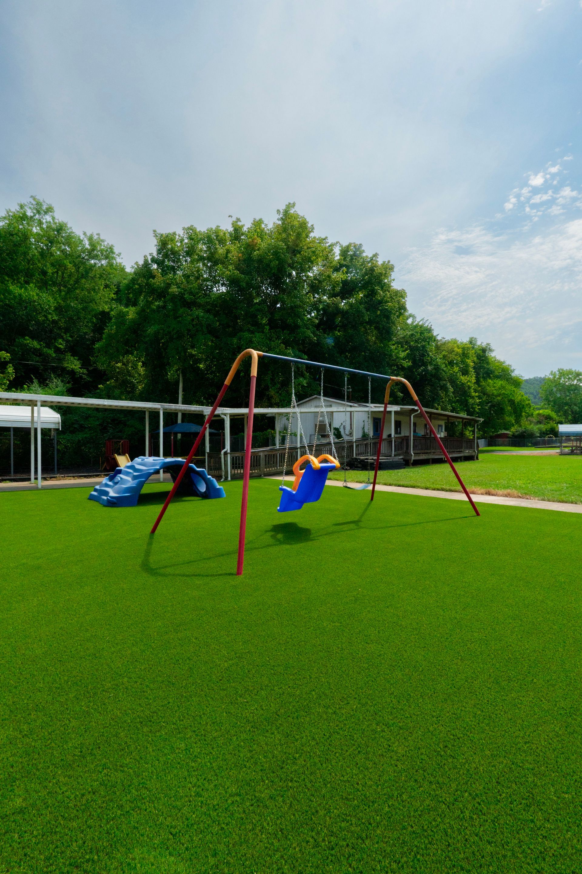 A playground with green turf, swings, and blue climbing structure. Trees and a building are in the background.