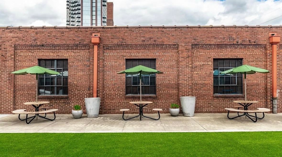 Outdoor dining area with picnic tables, green umbrellas, and a red brick wall.