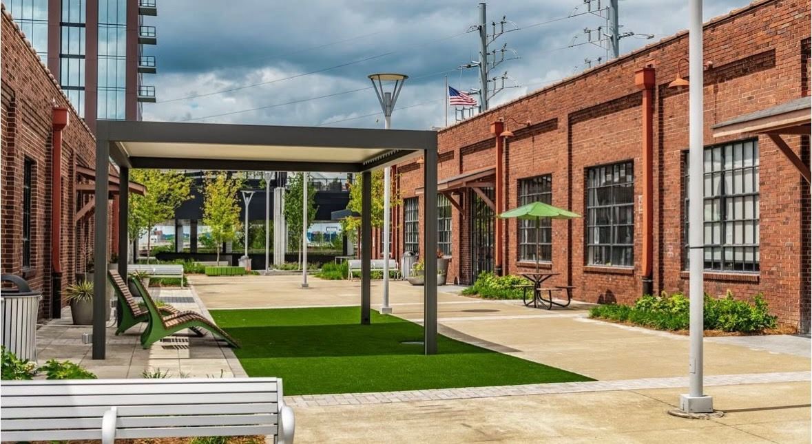 Courtyard with red brick buildings, pergola, benches, and green turf; overcast sky.
