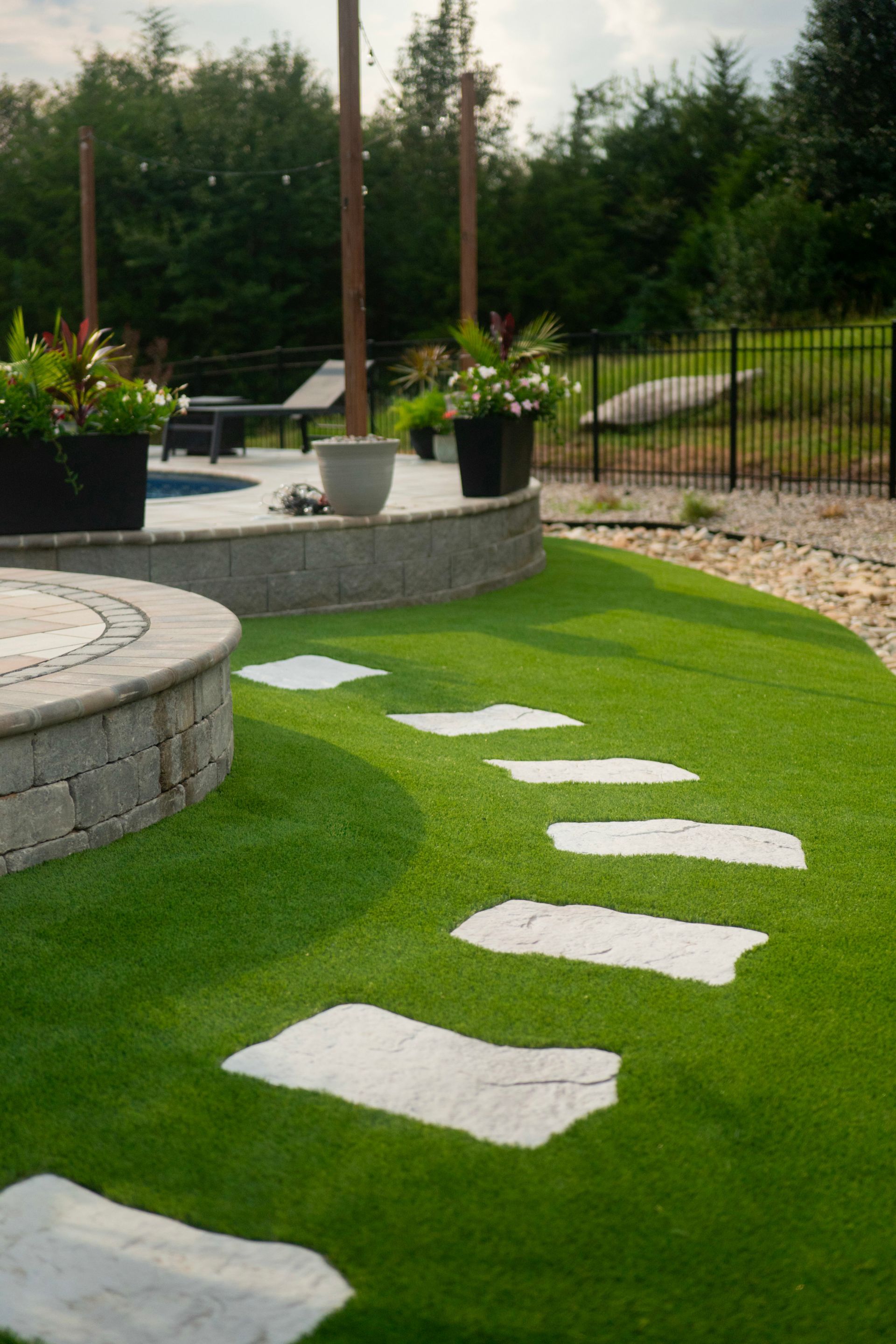 Stone pathway through artificial green grass leads to a patio area with potted plants, outdoor seating, and a dark fence.