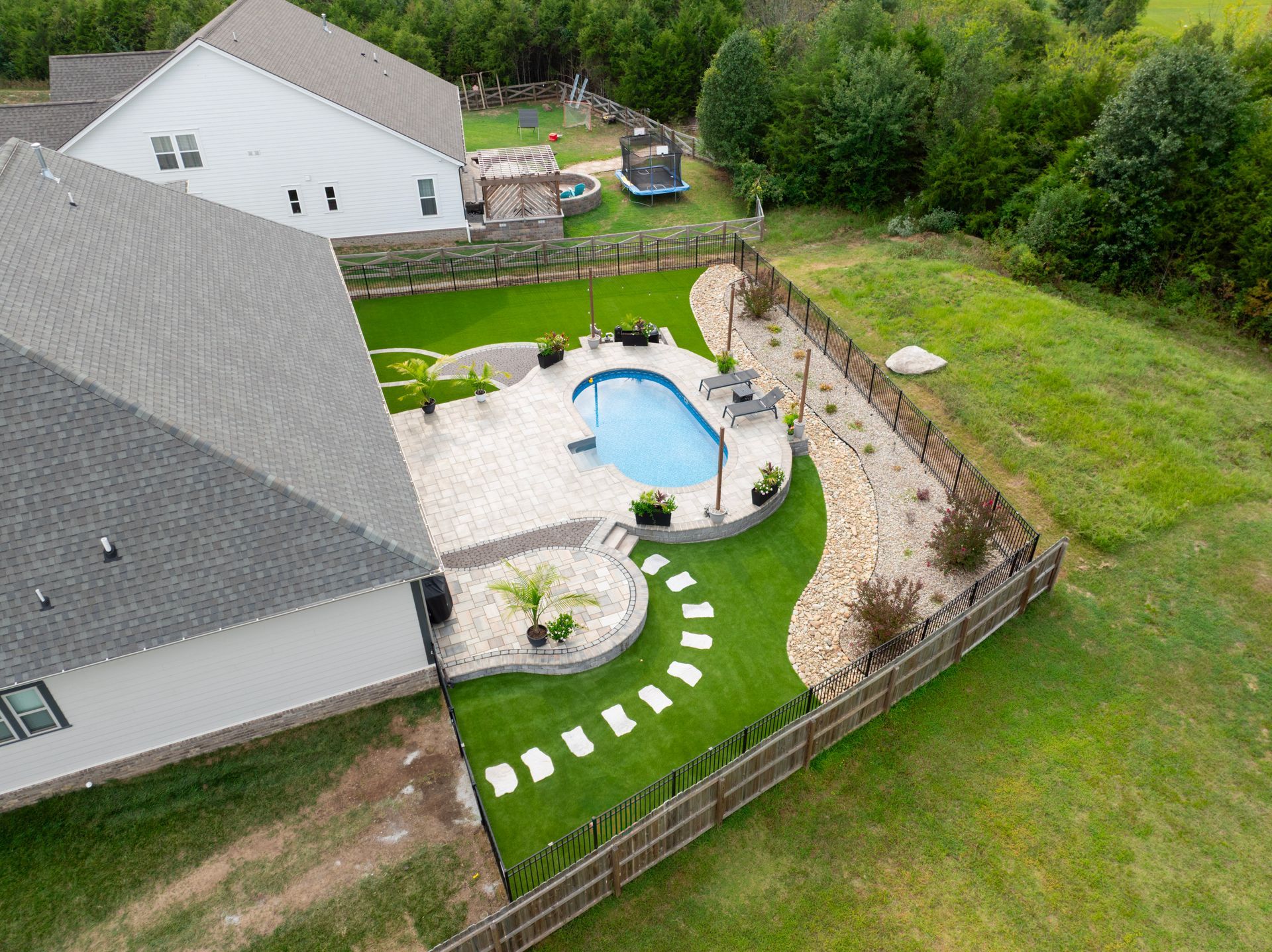Aerial view of a backyard with a pool, patio, landscaping, and a grassy area surrounded by a fence and a house.