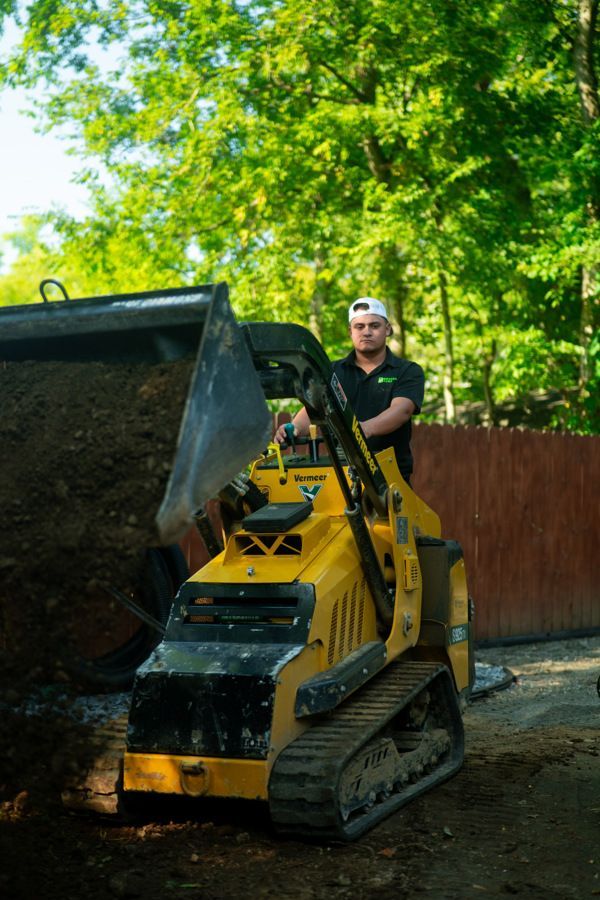 Man operating a yellow track skid steer, loading dirt. He wears a cap, black shirt, outdoors.