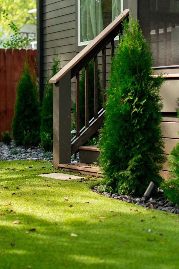 Green lawn and evergreens frame a dark-wood stair rail leading to a dark-sided house.