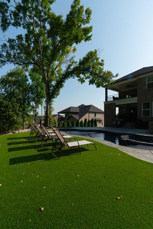 Lounge chairs on green lawn beside a pool, with a house and trees in the background on a sunny day.