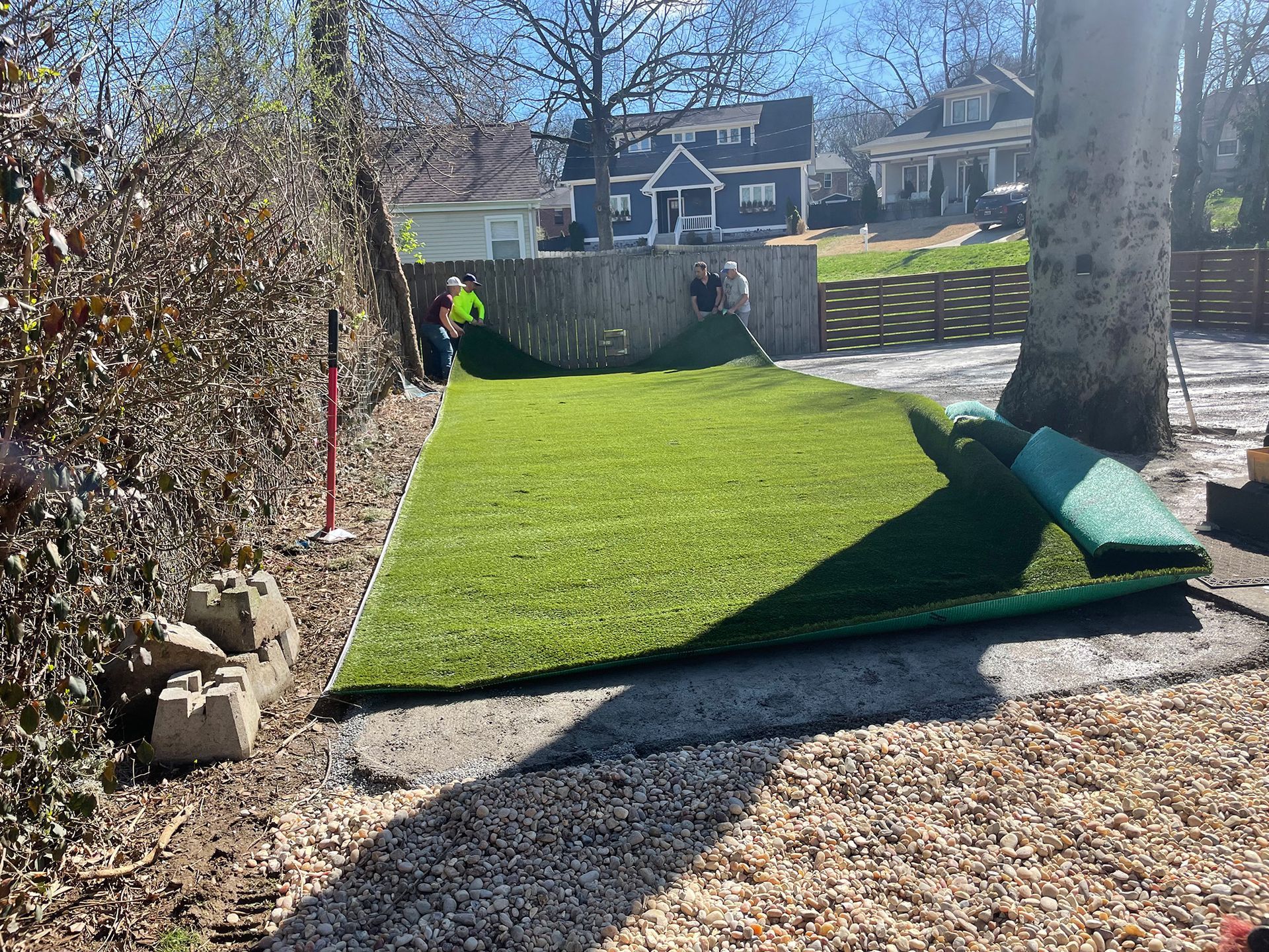 Workers installing artificial turf in a yard; green grass, fence, houses in the background.