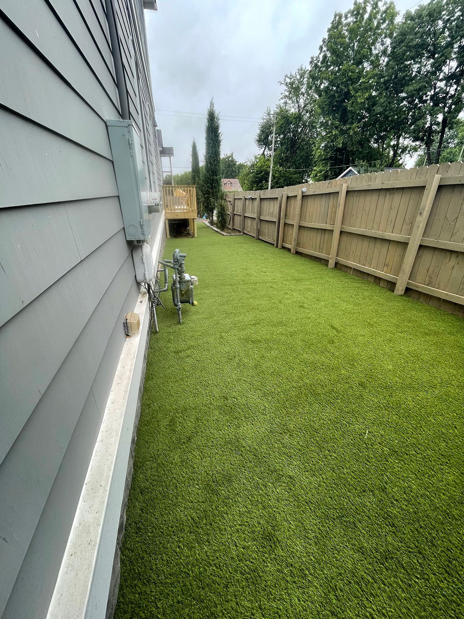 Side yard with artificial turf, wooden fence, and light blue house siding.