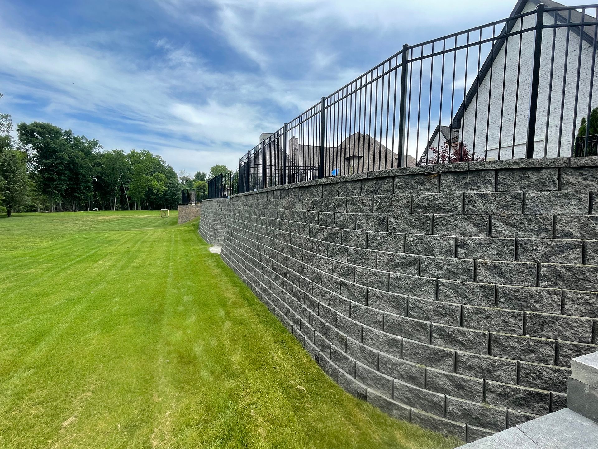 A tiered gray retaining wall with black fence atop, separates green grass lawn from a house under a blue sky.