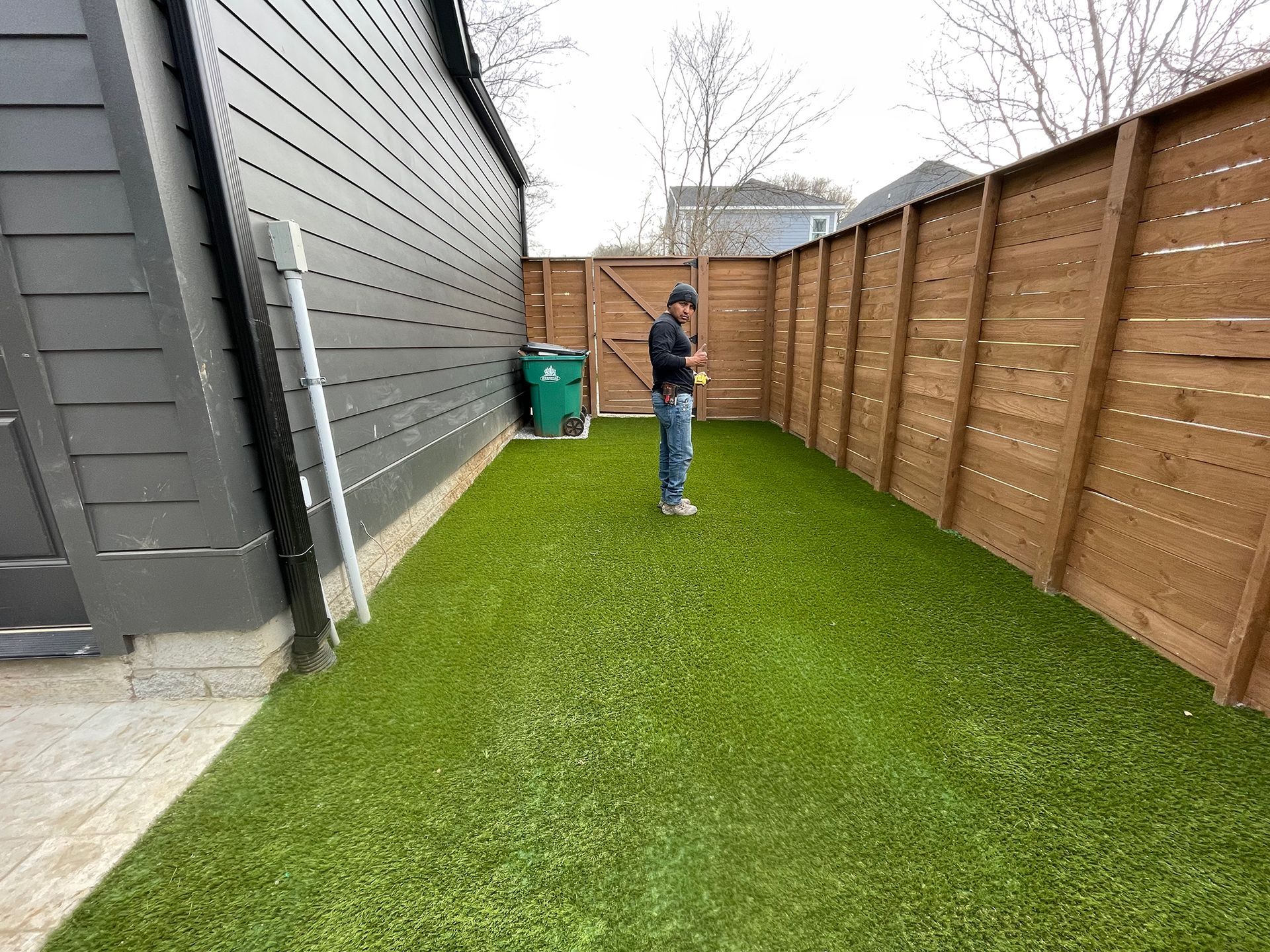 Person standing on green artificial turf in a narrow yard, next to a wooden fence and a dark-colored house.