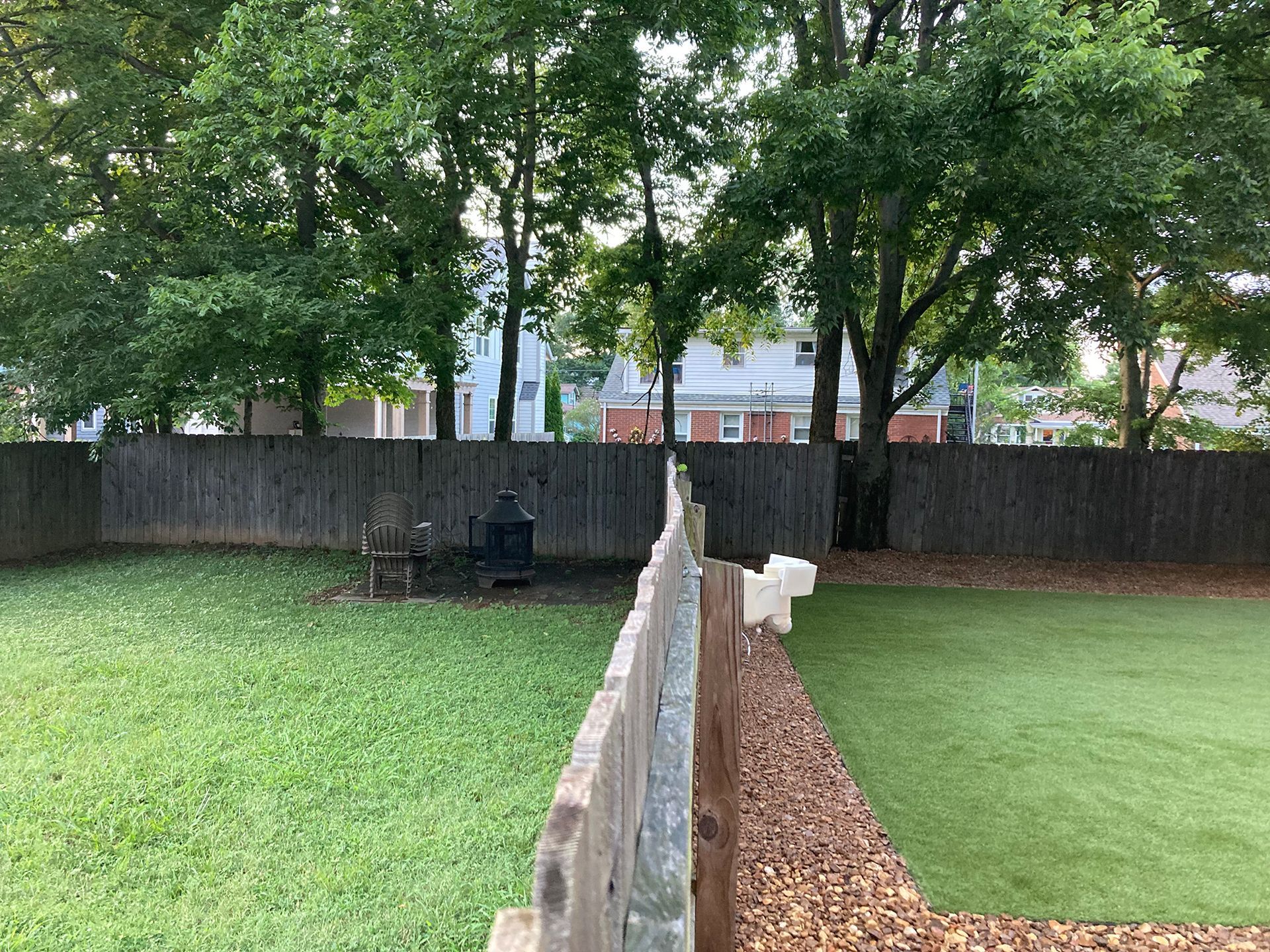 Backyard with a wooden fence, fire pit, chair, trees, grass, and a section of artificial turf.