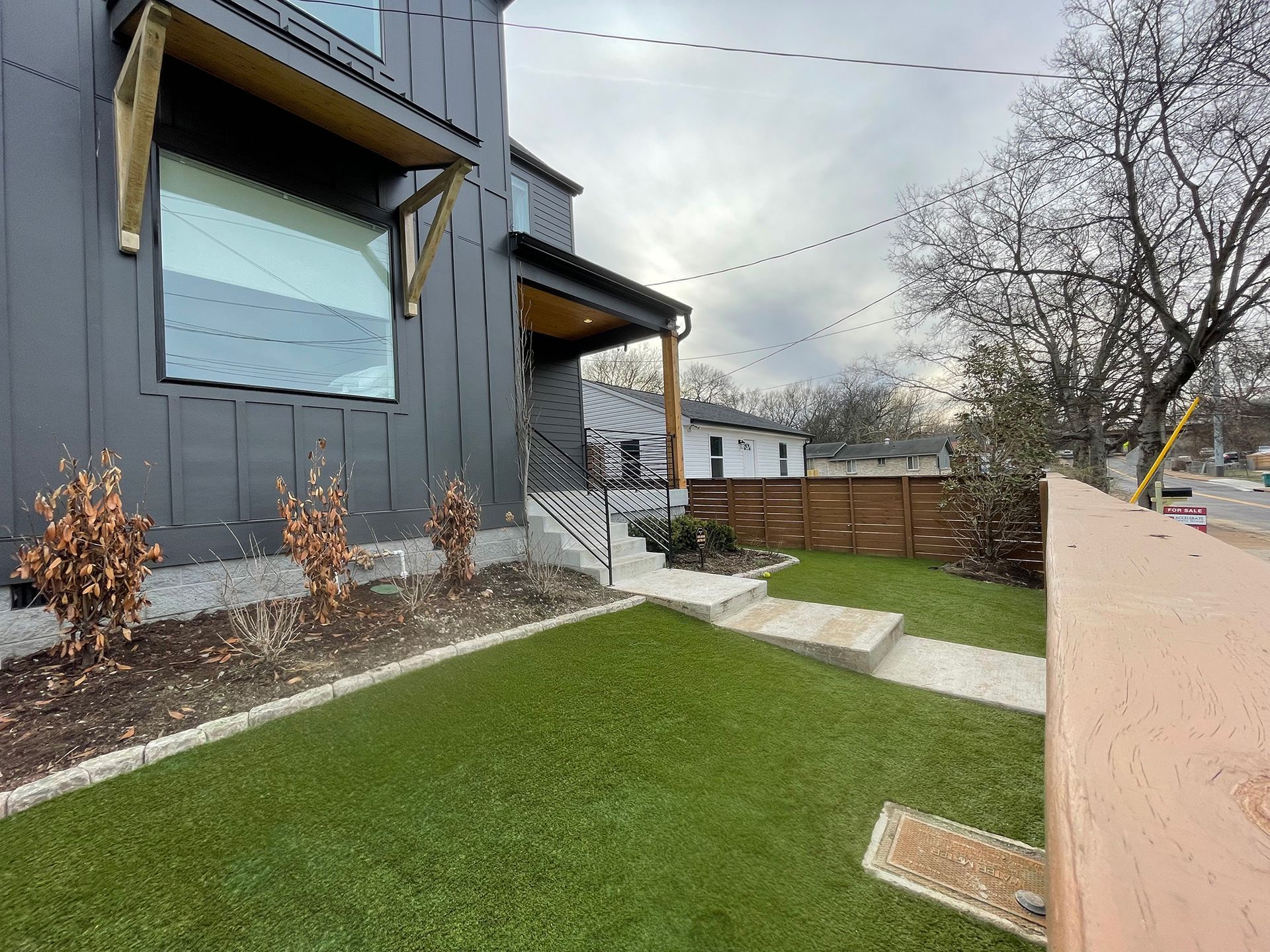 Modern gray house with wood accents, small front yard with artificial turf, cloudy sky.