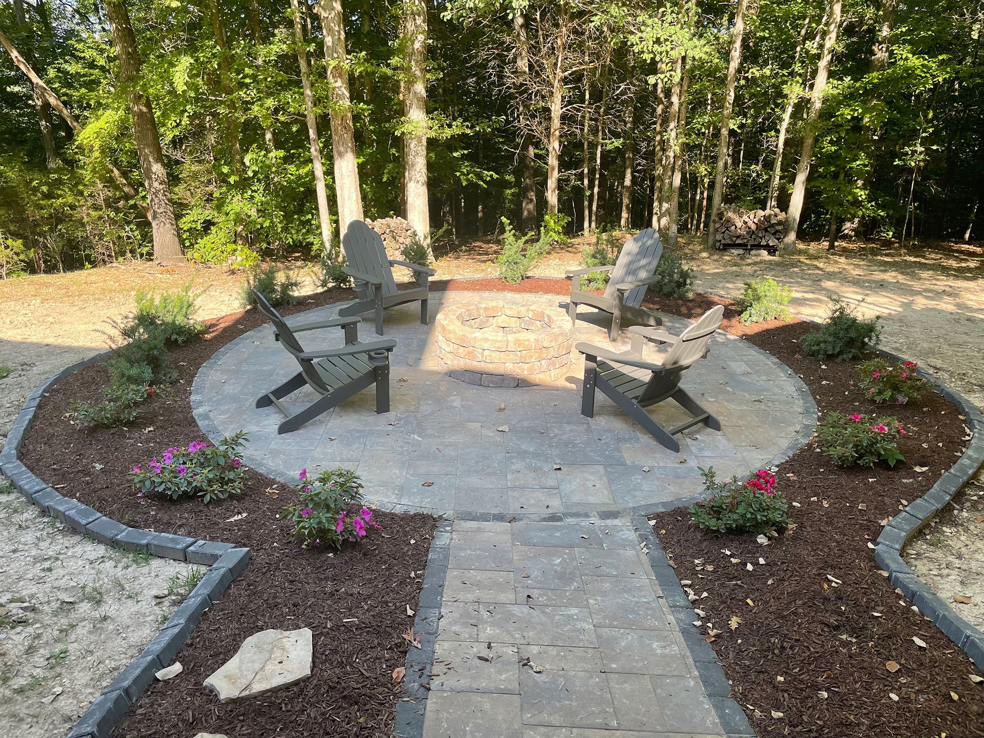 A circular patio with gray chairs, surrounded by plants and mulch, in a wooded area.