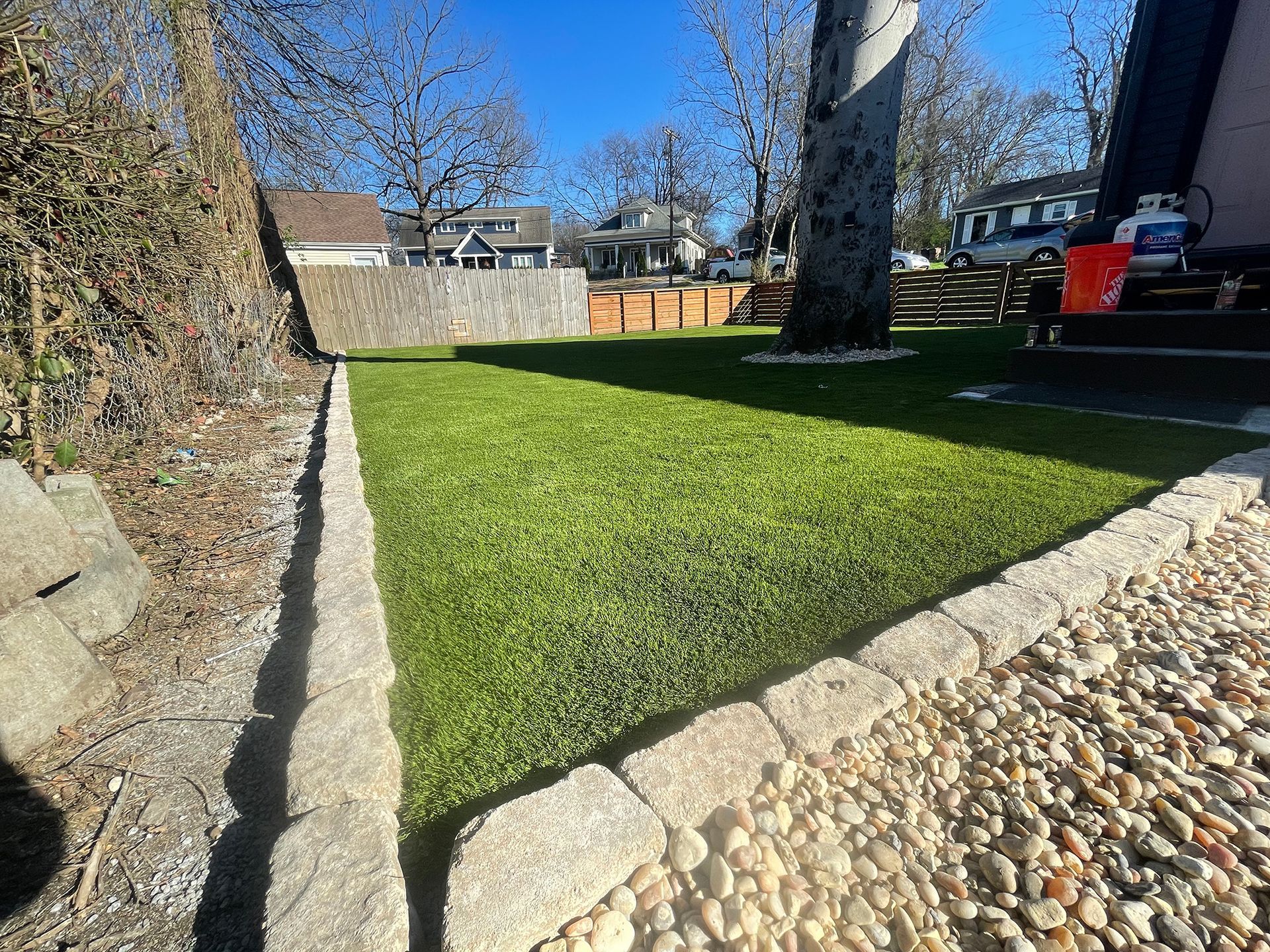 Well-manicured lawn bordered by stone and gravel, trees in background, clear blue sky.