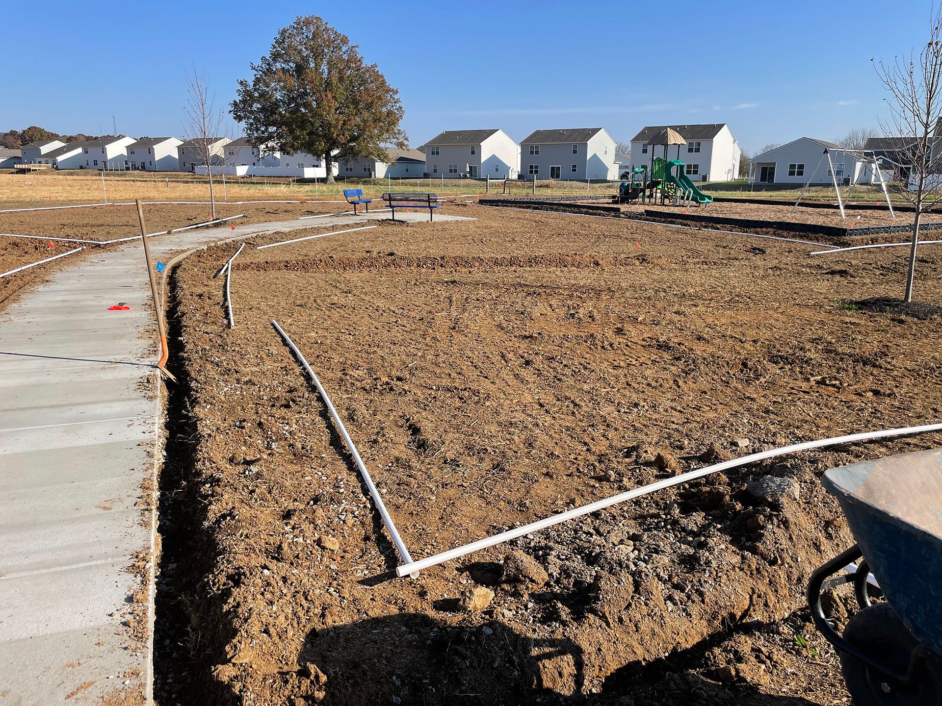 Construction site: a paved path with irrigation lines in dirt near a playground, houses in background.