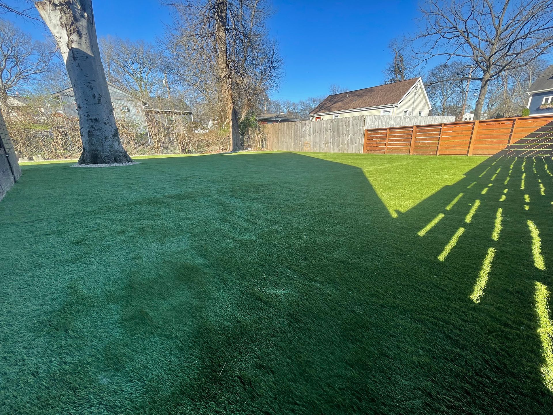 Green artificial turf backyard with fence, trees, and house under blue sky.
