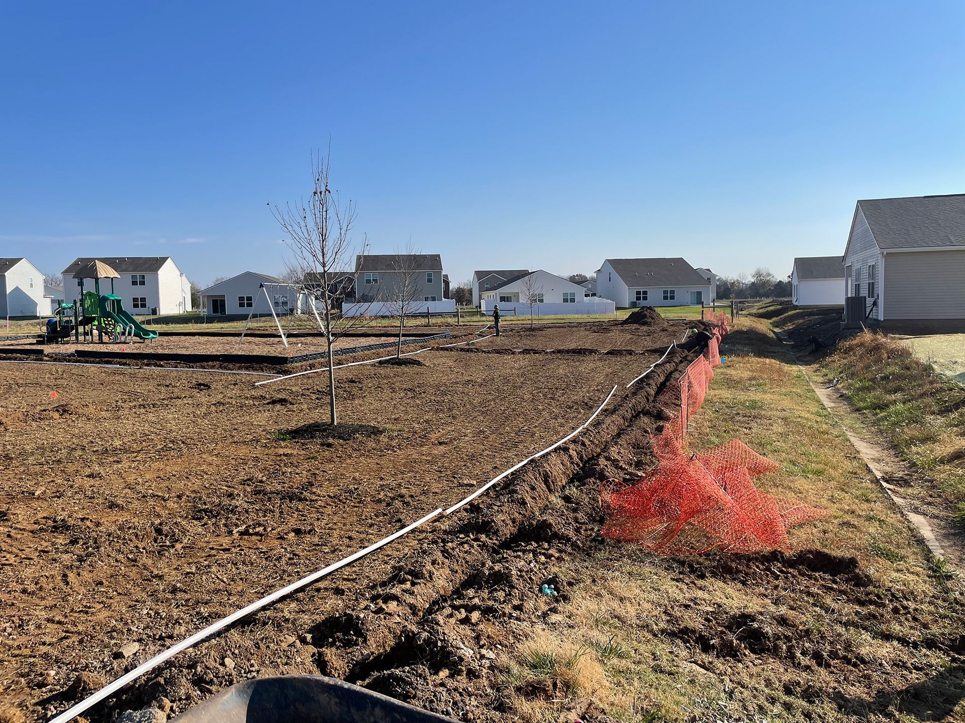 Construction site with dirt, white pipe, orange barrier, houses in background.