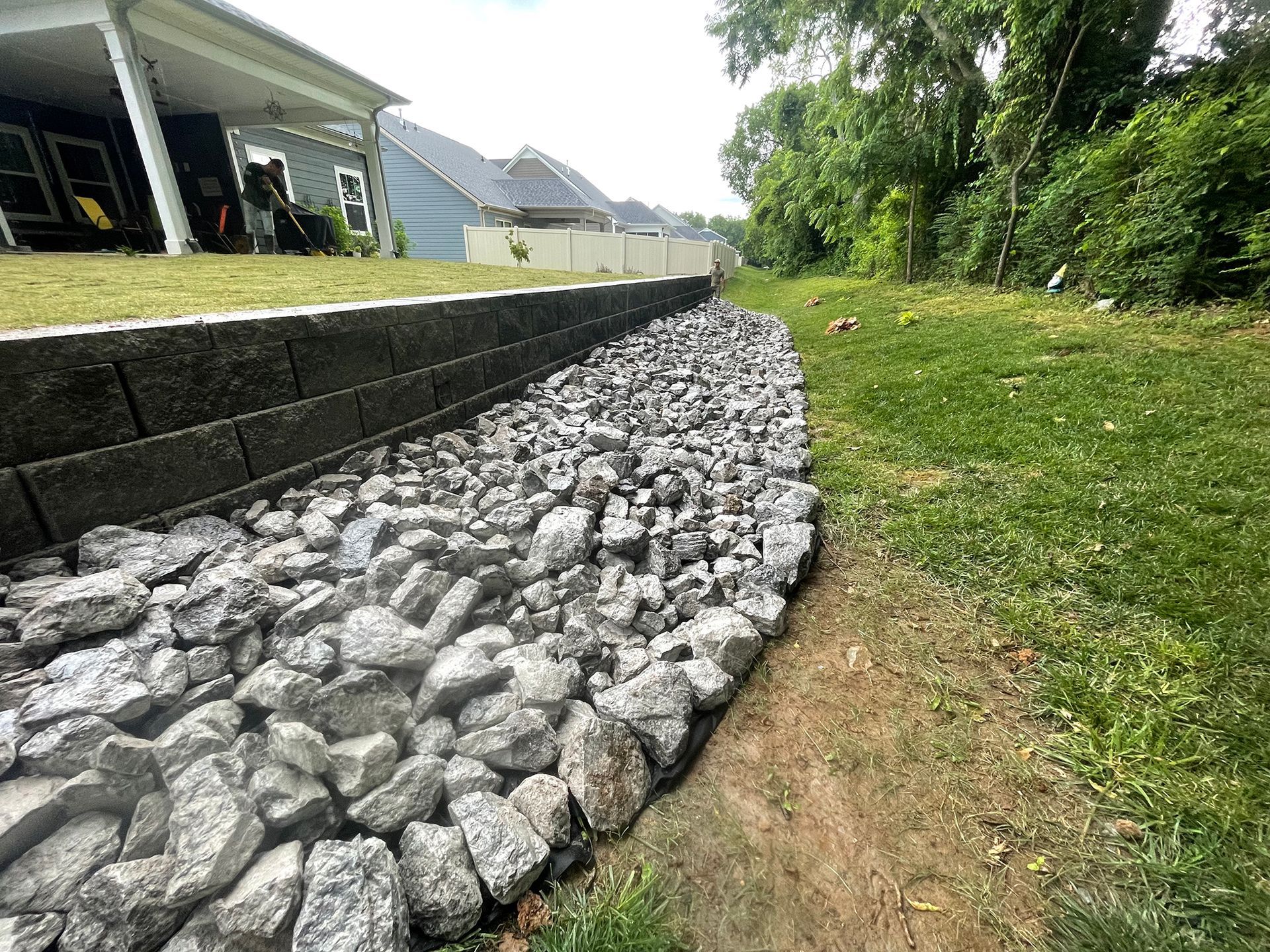 A stone-filled drainage channel runs along a grassy bank beside a retaining wall, next to a house.