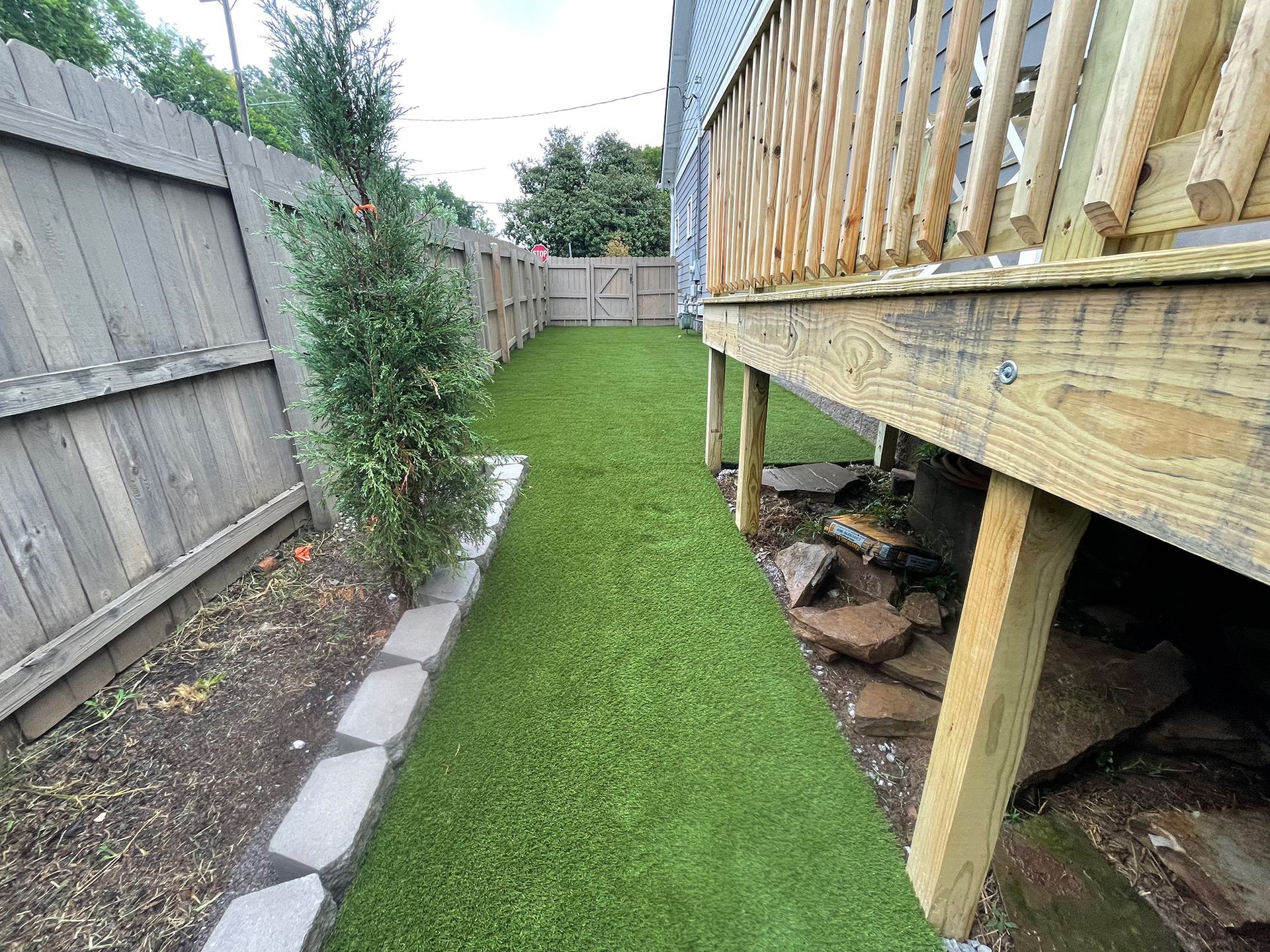 Narrow backyard with green turf, wooden deck, gray fence, and small tree.