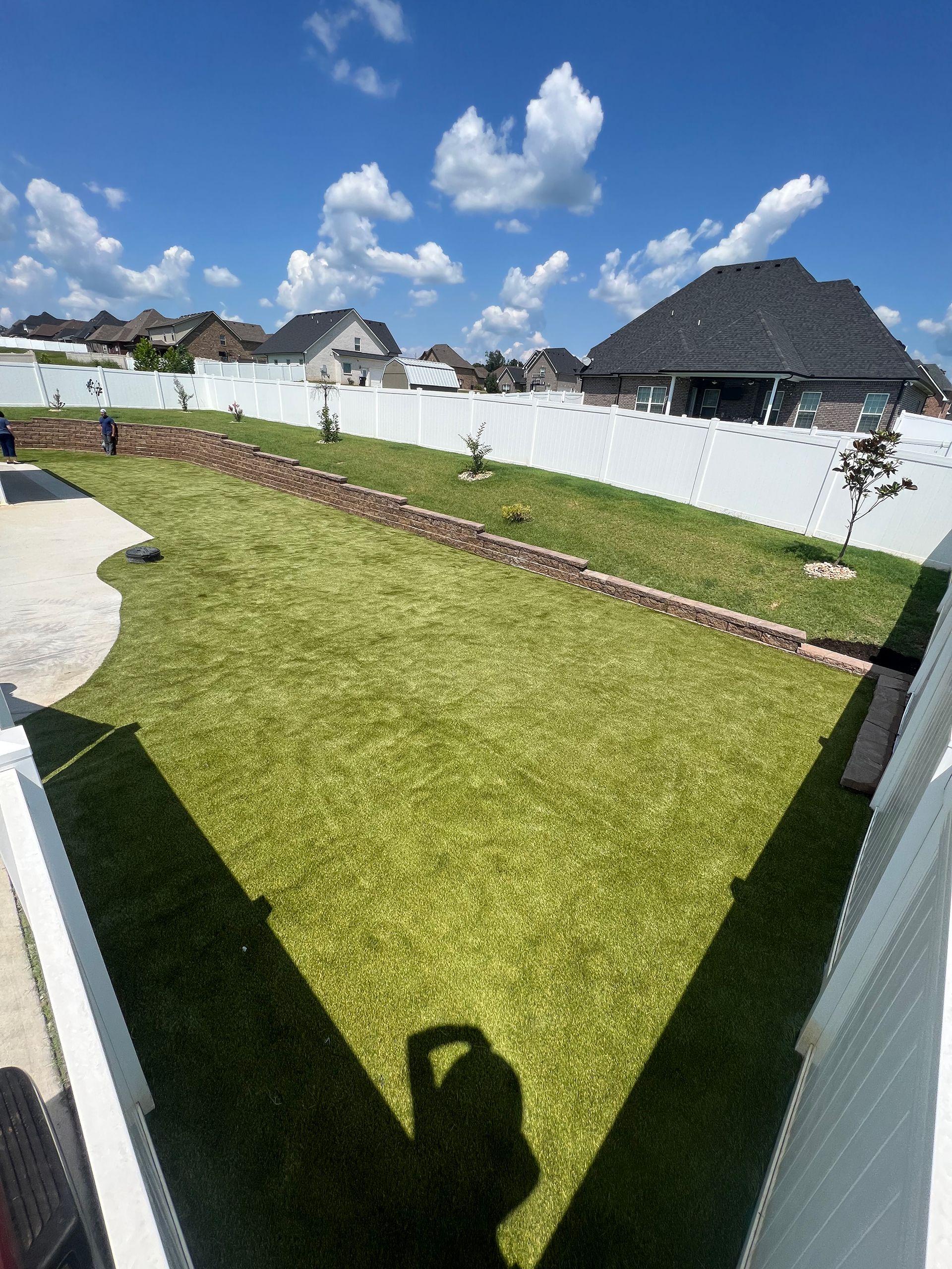 Green lawn in a fenced backyard, terraced with a retaining wall. Sunny day with blue sky and clouds.