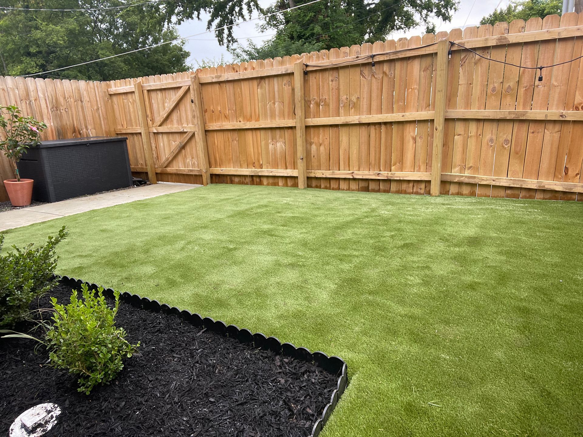 A backyard with green artificial turf, black mulch border, wooden fence, and storage bin.