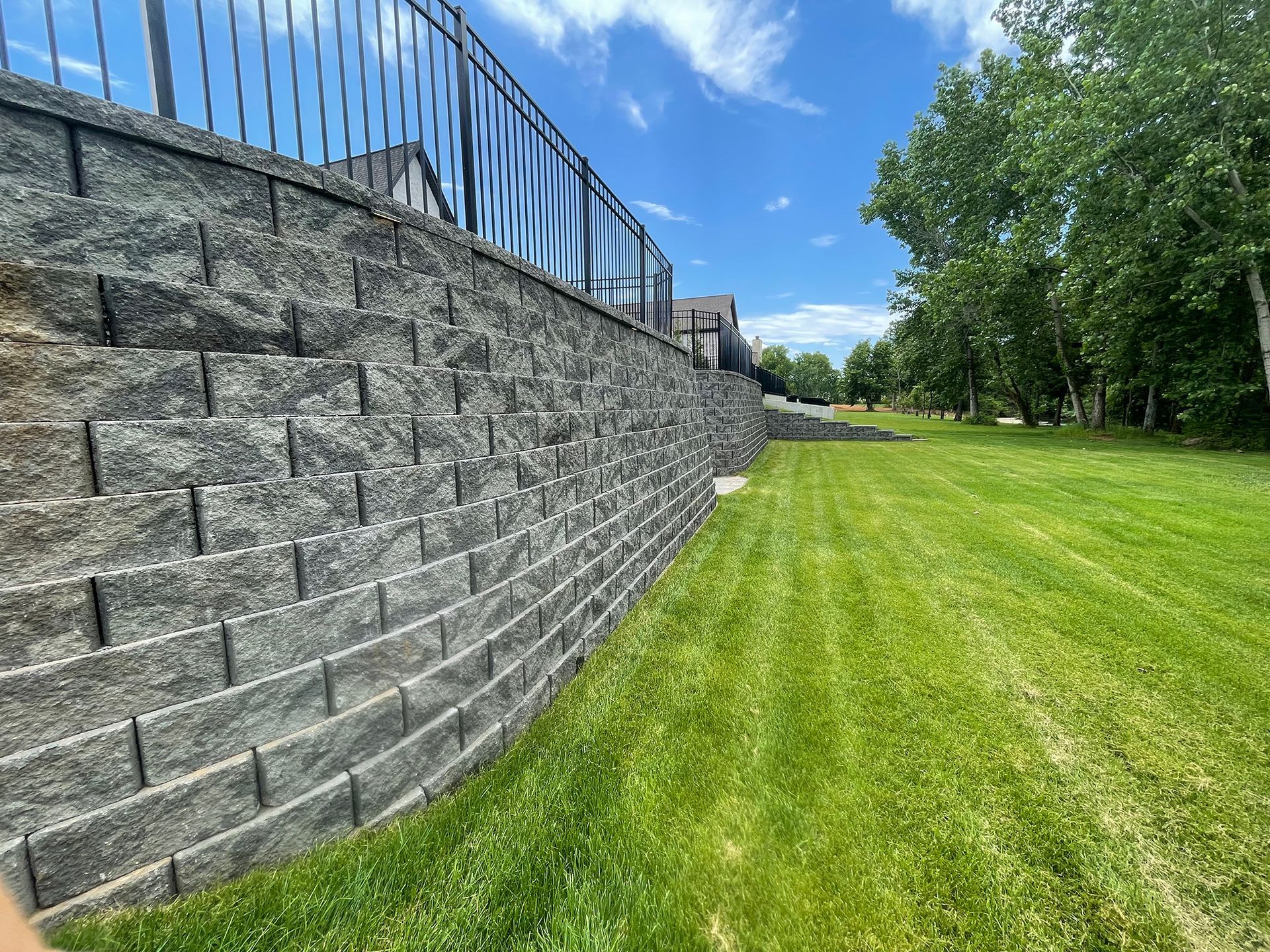 Stone retaining wall with black fence, green grass, and trees under a blue sky.