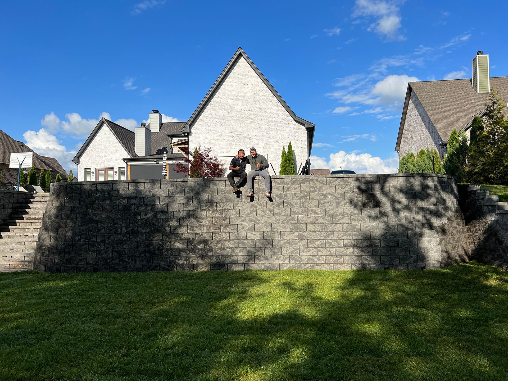 Two people sit on a retaining wall in front of a house on a sunny day.