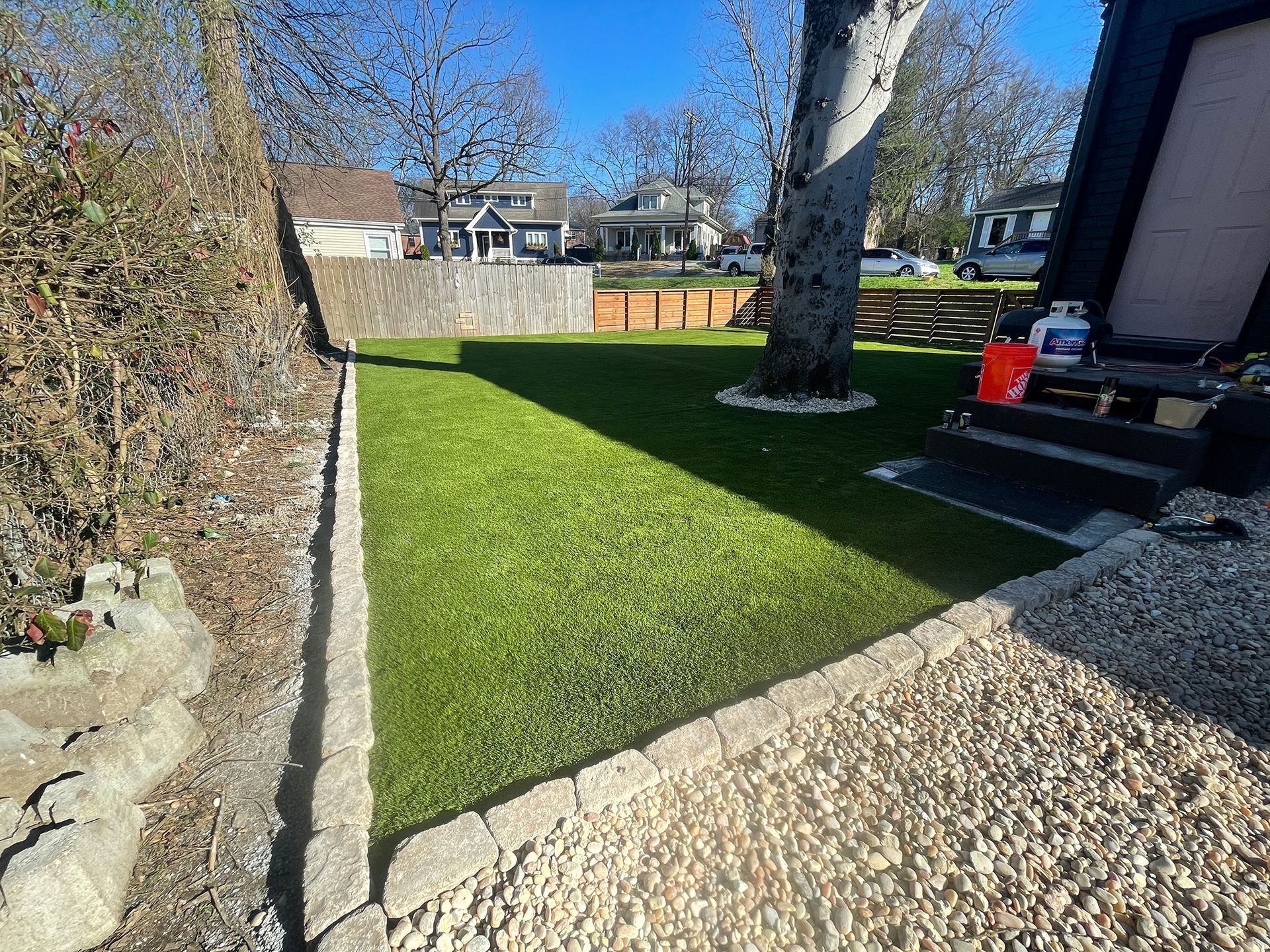 Green lawn bordered by stone edging, gravel, and a retaining wall. Houses visible in background.