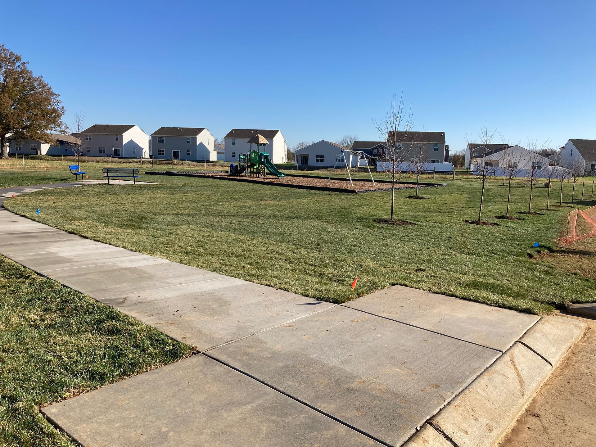 A park with a playground, bench, and houses on a sunny day. Concrete path in the foreground.