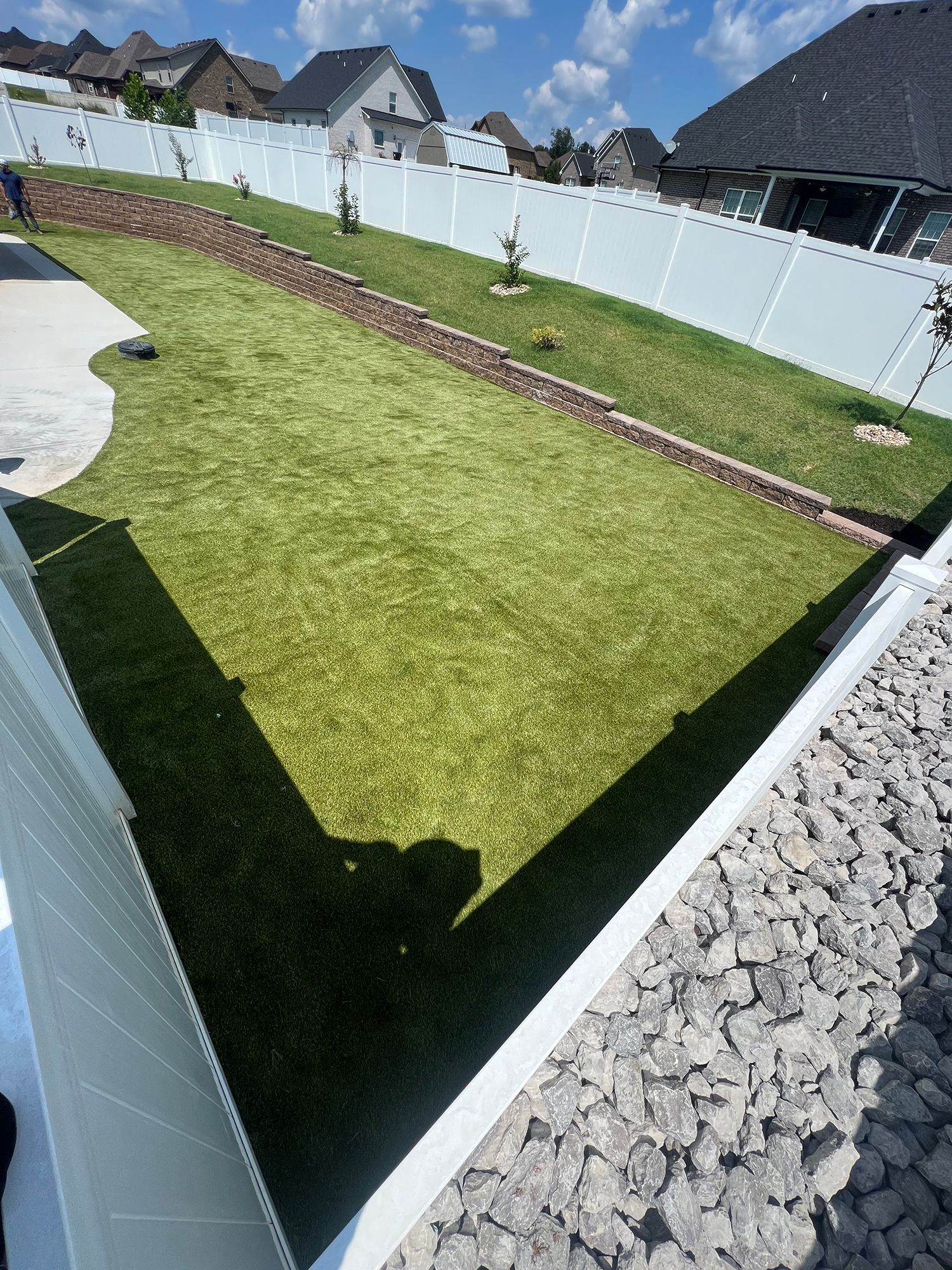Green lawn bordered by white fence and rock, on a sunny day.