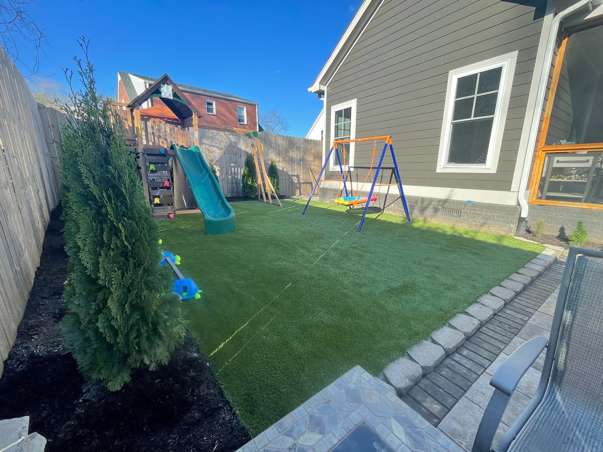 A backyard with a playground, artificial turf, and a house with gray siding on a sunny day.
