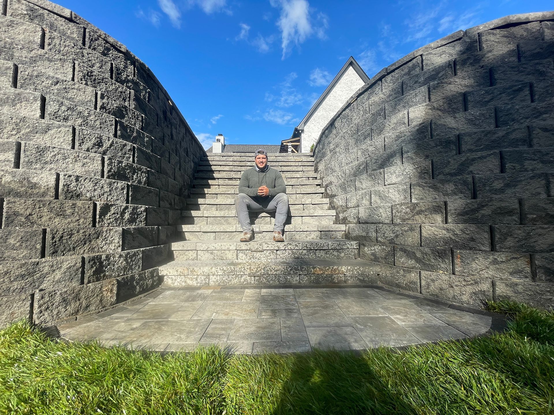 Man sits on stone steps between two textured stone walls, blue sky overhead.