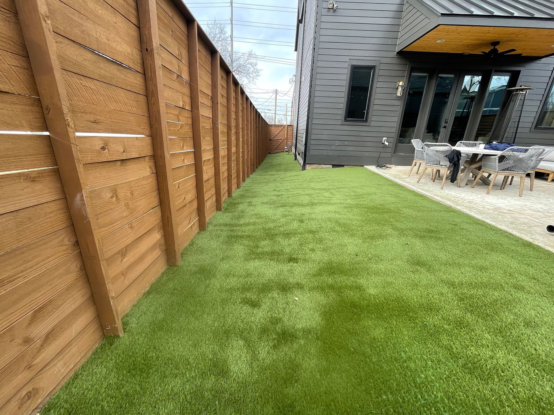 Lush green artificial turf lawn between a brown wooden fence and modern gray house.