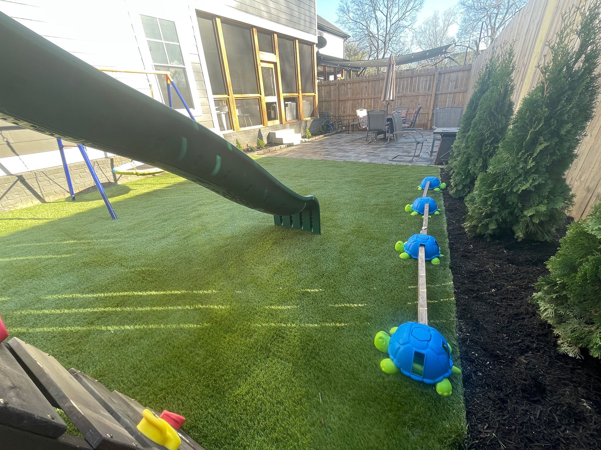 A backyard with green turf, a slide, and a turtle-themed climbing toy. Thuja shrubs line the wooden fence.