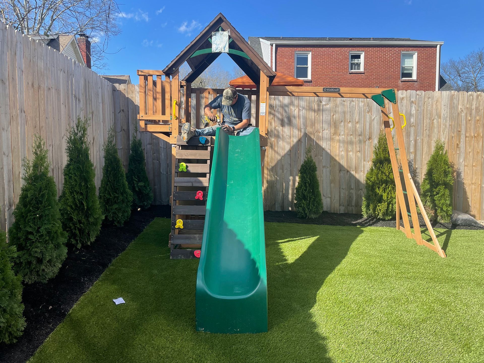 Child plays on a wooden playset with slide and climbing wall in a backyard with a fence and green grass.