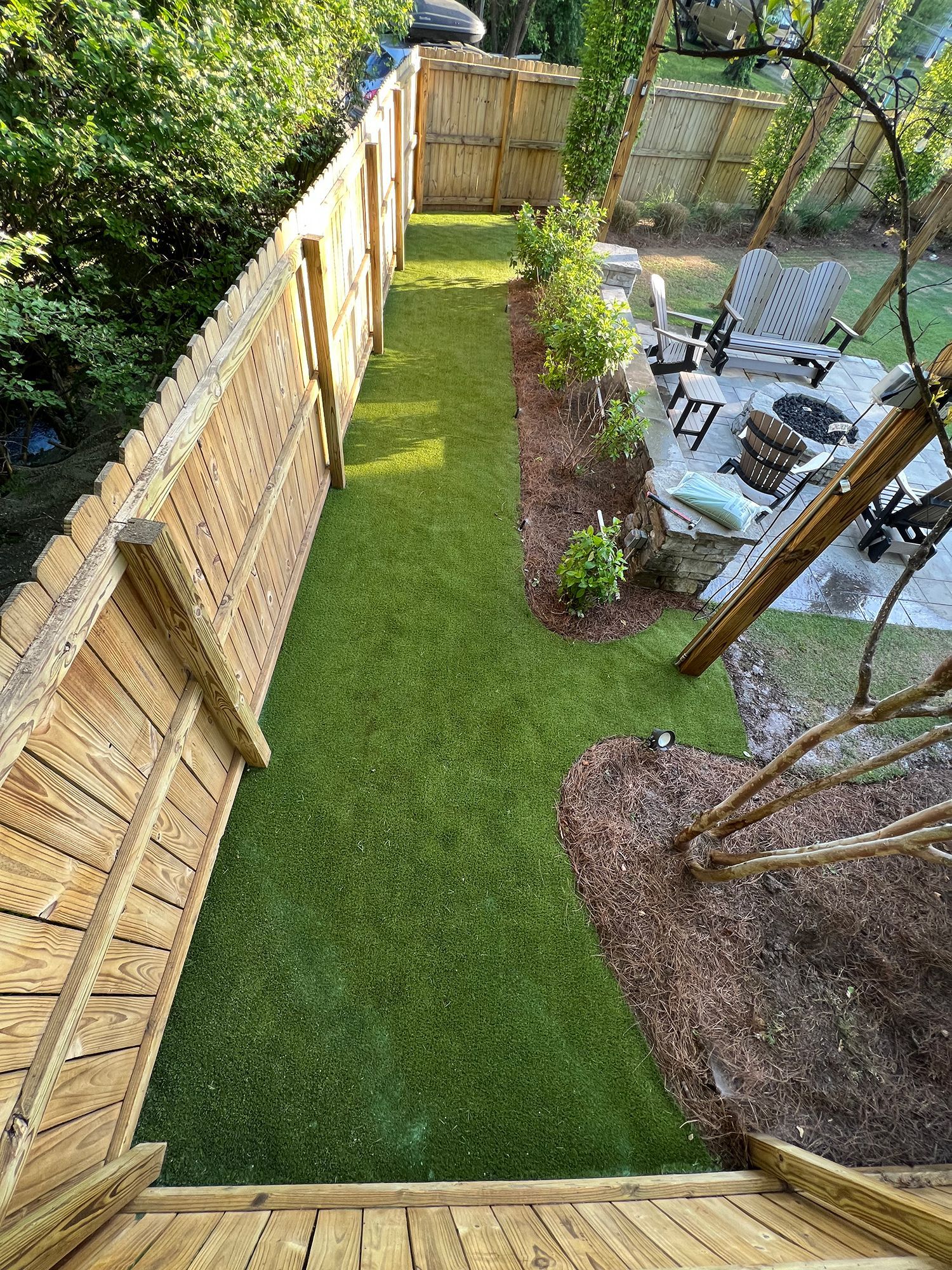 Long, narrow backyard with green grass path between wooden fences, leading to a patio with furniture.