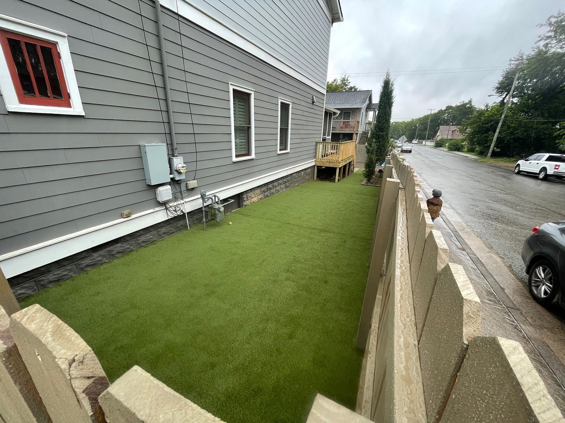 Side view of a grey house with a green turf yard, wooden fence, and street.