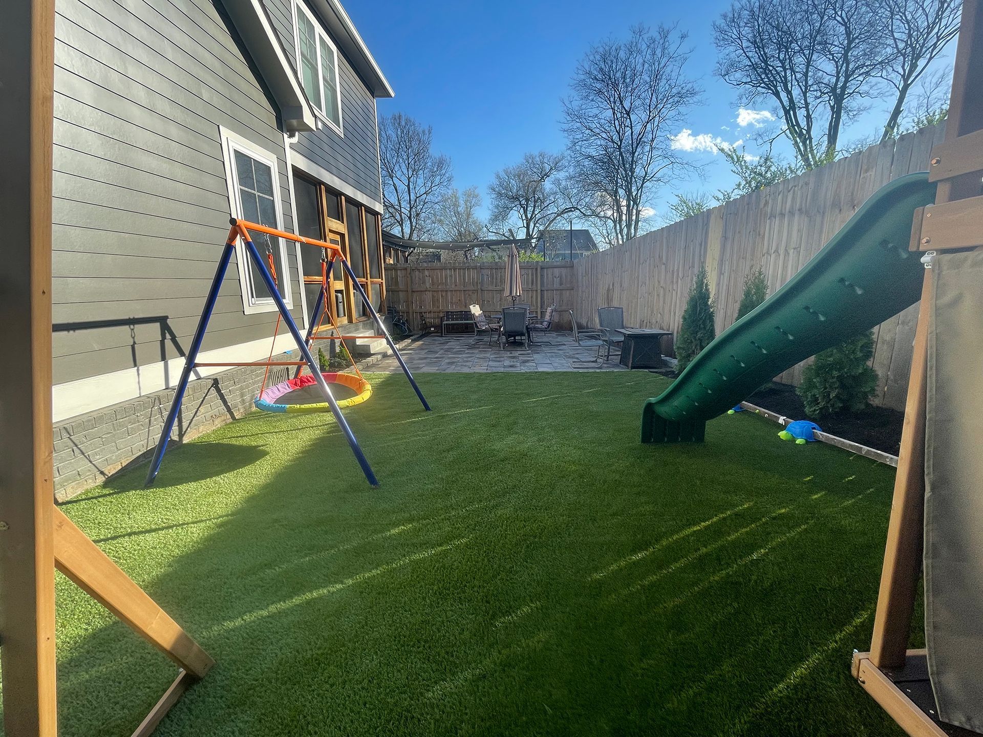 Backyard with swing set, slide, and artificial grass. Gray house and bamboo fence in the background.