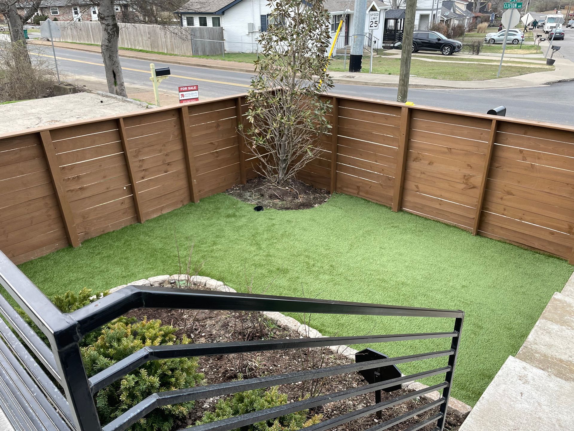 A backyard with brown fence, green turf, a small tree, and black railing. Road and houses in the background.