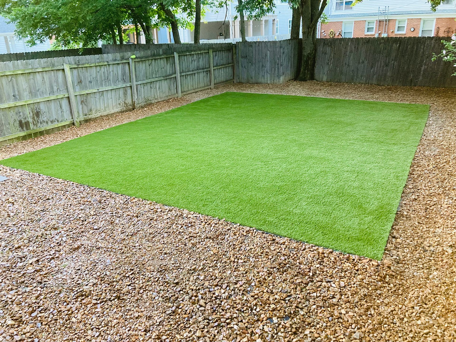 Green artificial turf in a gravel yard, bordered by a wooden fence.